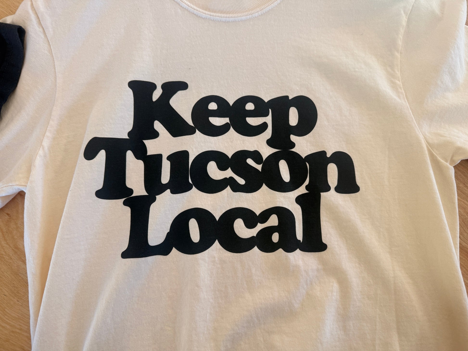 White t-shirt with 'Keep Tucson Local' text in black on a wooden surface
