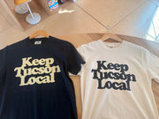 Two t-shirts with 'Keep Tucson Local' text on hangers on a wooden floor.