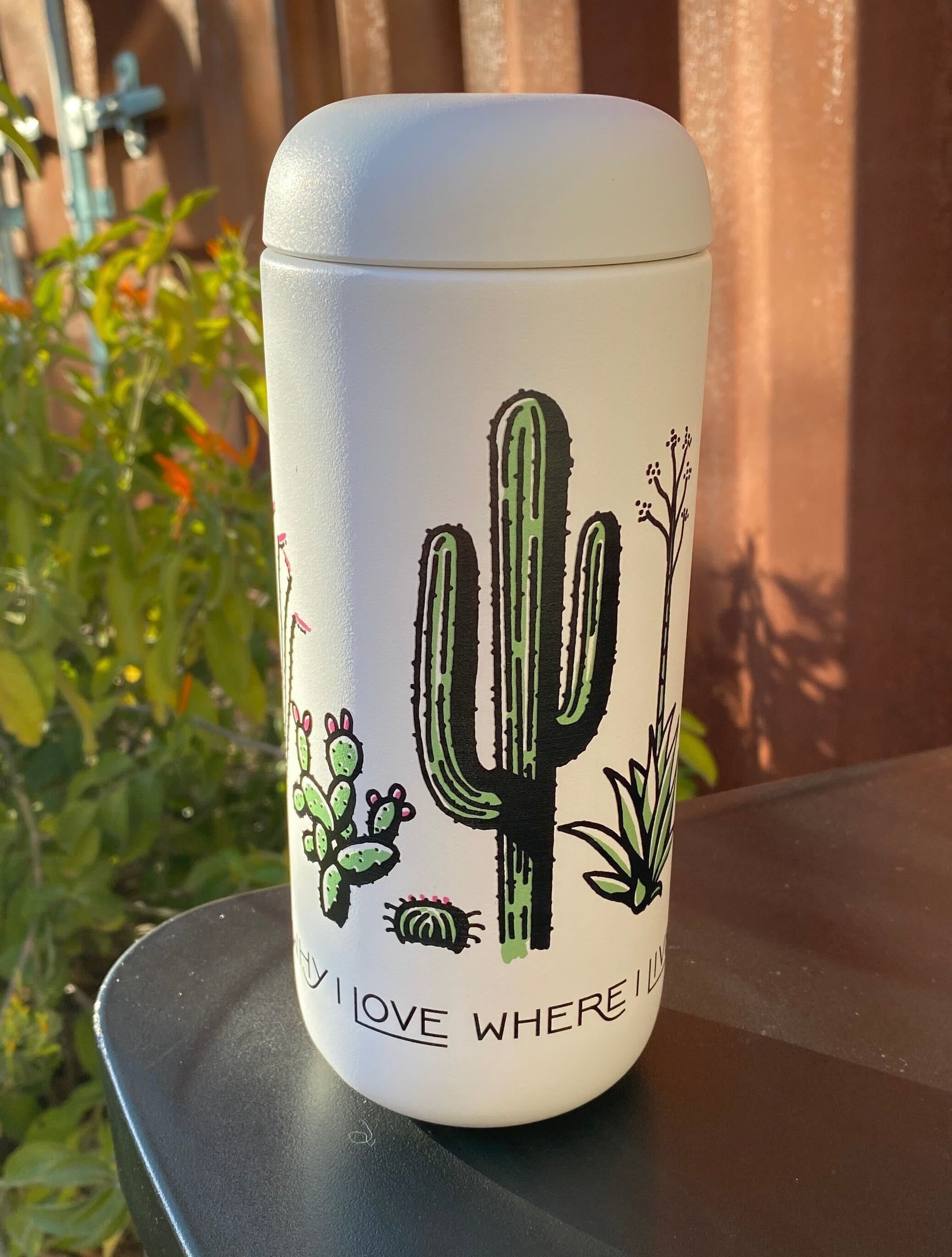 A white coffee tumbler with a cactus design, a lid, and text on the front, placed on a table outdoors.